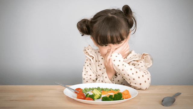 Child with a displeased expression, covering her face while sitting at a table with a plate of vegetables.