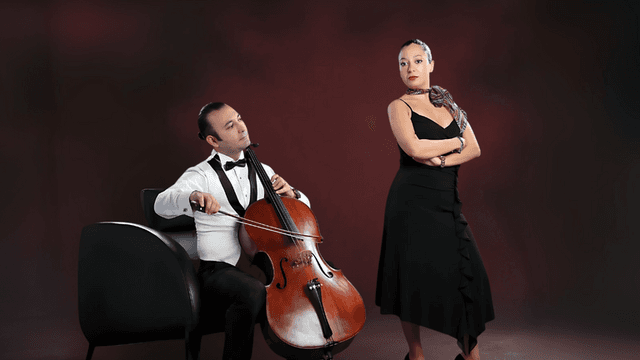 Man playing cello while woman in black dress stands beside him on stage.