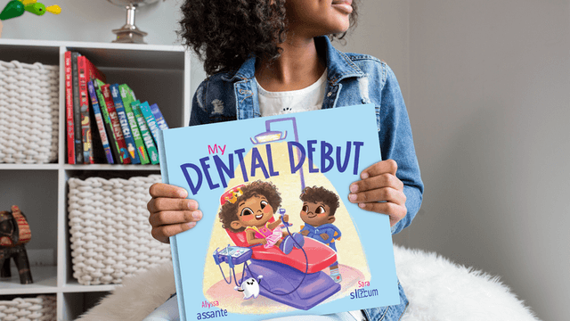 A child holds the book My Dental Debut, featuring two animated characters at a dental appointment.