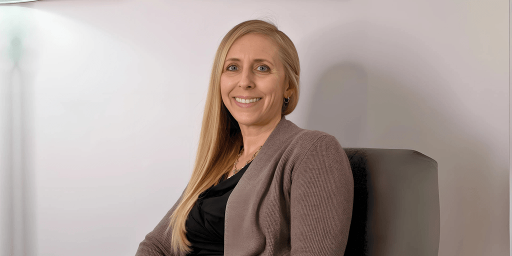 Smiling woman with long blonde hair, wearing a brown cardigan and a black top, sitting comfortably in a chair with soft lighting and a simple background.