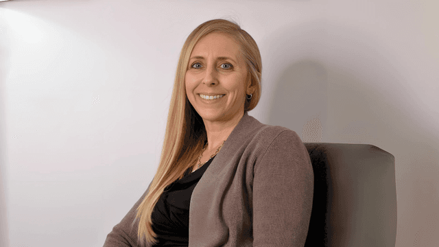 Smiling woman with long blonde hair, wearing a brown cardigan and a black top, sitting comfortably in a chair with soft lighting and a simple background.