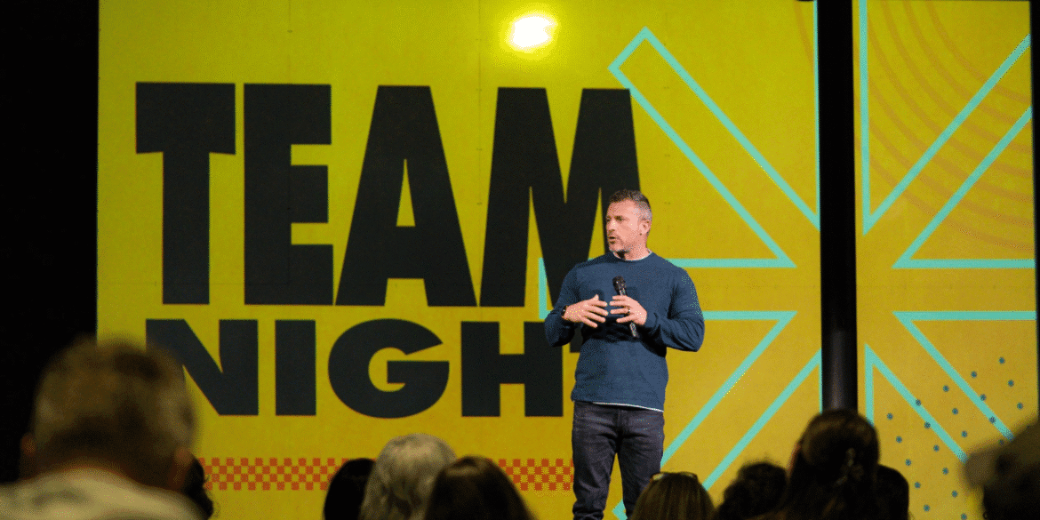 Speaker presenting at a Team Night event, with a vibrant yellow backdrop and bold 'TEAM NIGHT' text. The speaker is engaging with the audience, emphasizing teamwork and motivation. The image captures a professional and energetic atmosphere at a leadership or team-building gathering.