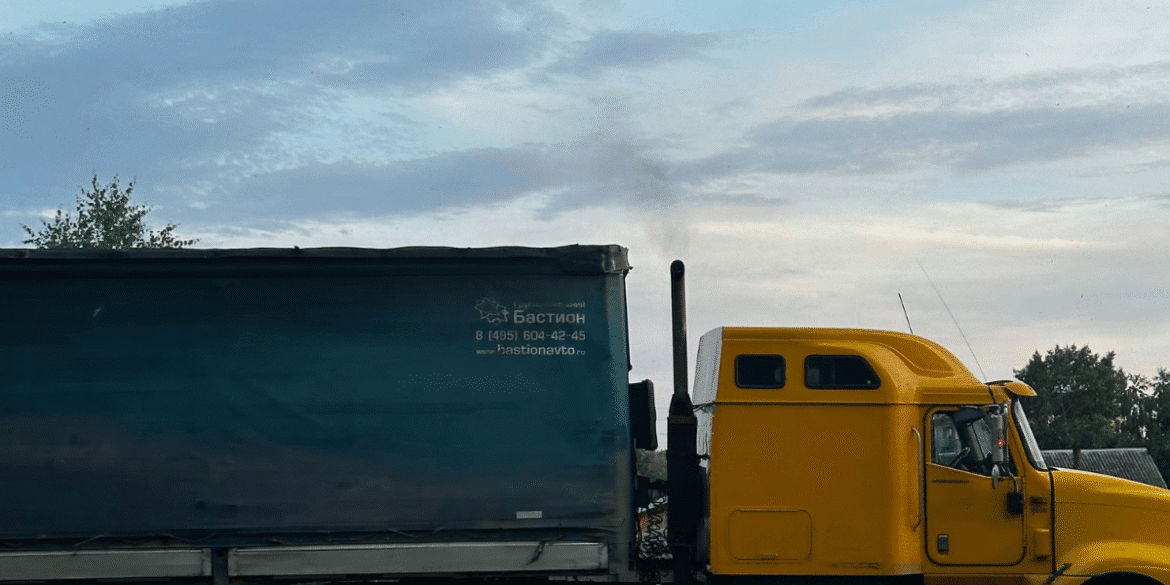 This image shows a yellow truck with a large, blue tarped trailer, with visible smoke coming from its exhaust pipe. The truck is marked with the text "Бастион" and includes a contact number and website, suggesting it's related to a transportation or logistics company. The truck is positioned in a rural or industrial setting, with a clear sky and some greenery in the background. The view focuses on the side of the truck, with the exhaust pipe releasing smoke into the air.