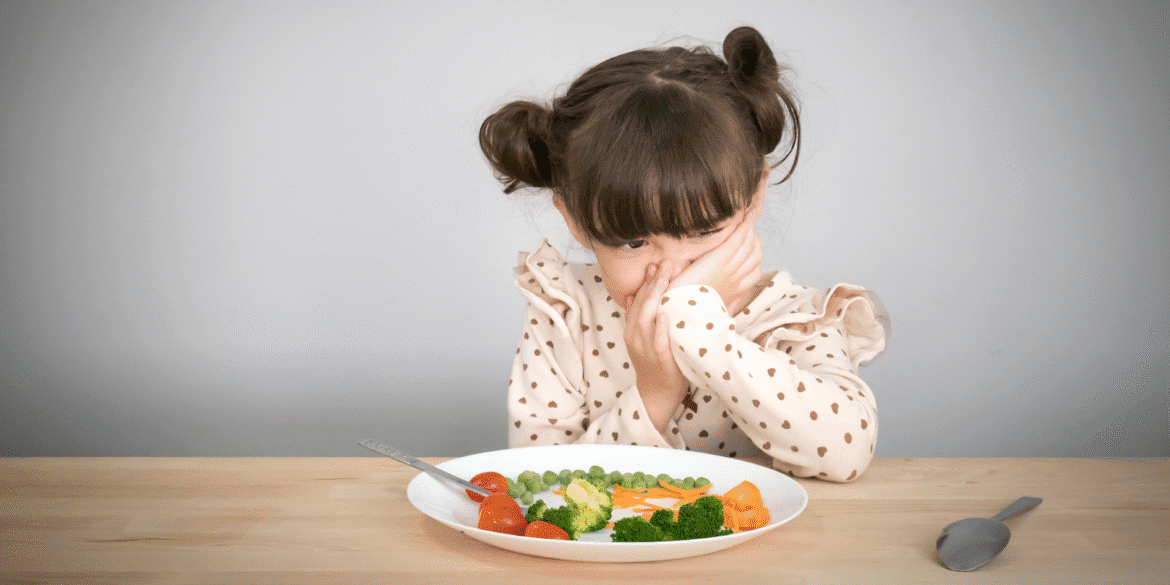 Child with a displeased expression, covering her face while sitting at a table with a plate of vegetables.
