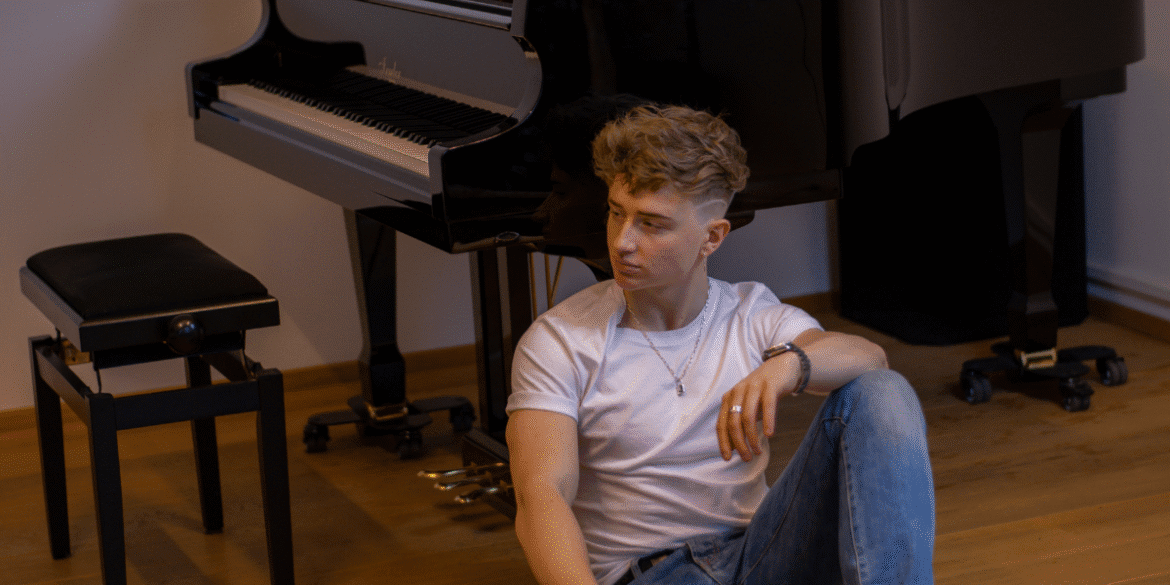 "Young male musician sitting on the floor next to a grand piano, wearing a white t-shirt and jeans, looking contemplative with his hand resting on his knee, exuding a relaxed and thoughtful atmosphere."