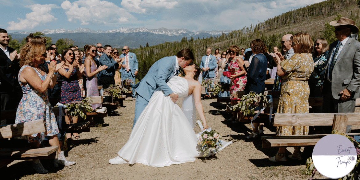 A wedding couple sharing a kiss during their outdoor ceremony, surrounded by guests cheering, with a stunning mountain backdrop.