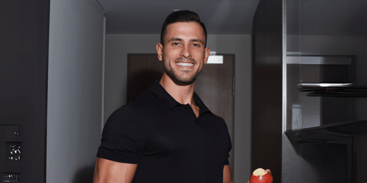 "Man in a black polo shirt holding a red apple, smiling in a modern kitchen setting with shelves in the background."