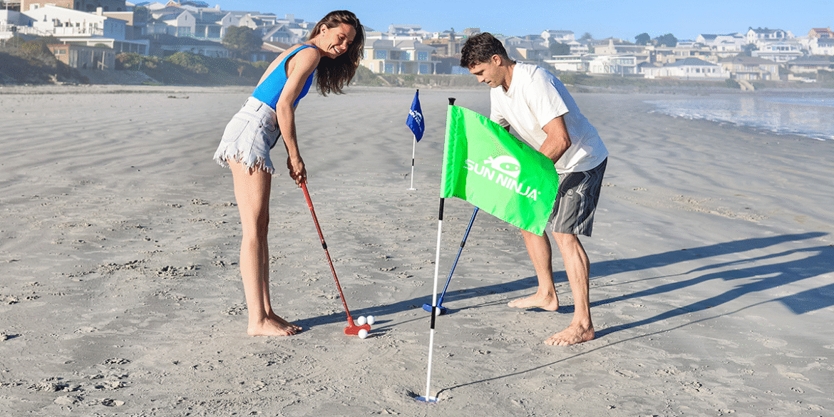 "Couple enjoying beach golf with colorful flags and golf clubs, one of them playing with a red club and the other adjusting the green flag."