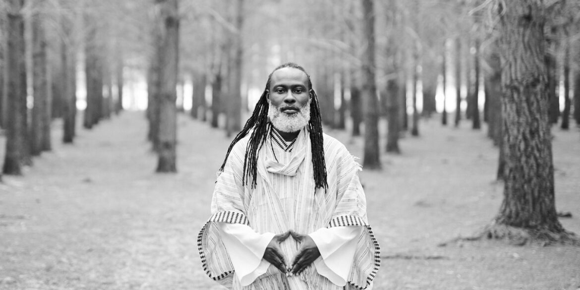 Black-and-white photo of SPEARHEART standing in a forest, wearing traditional patterned robe, looking directly at the camera.