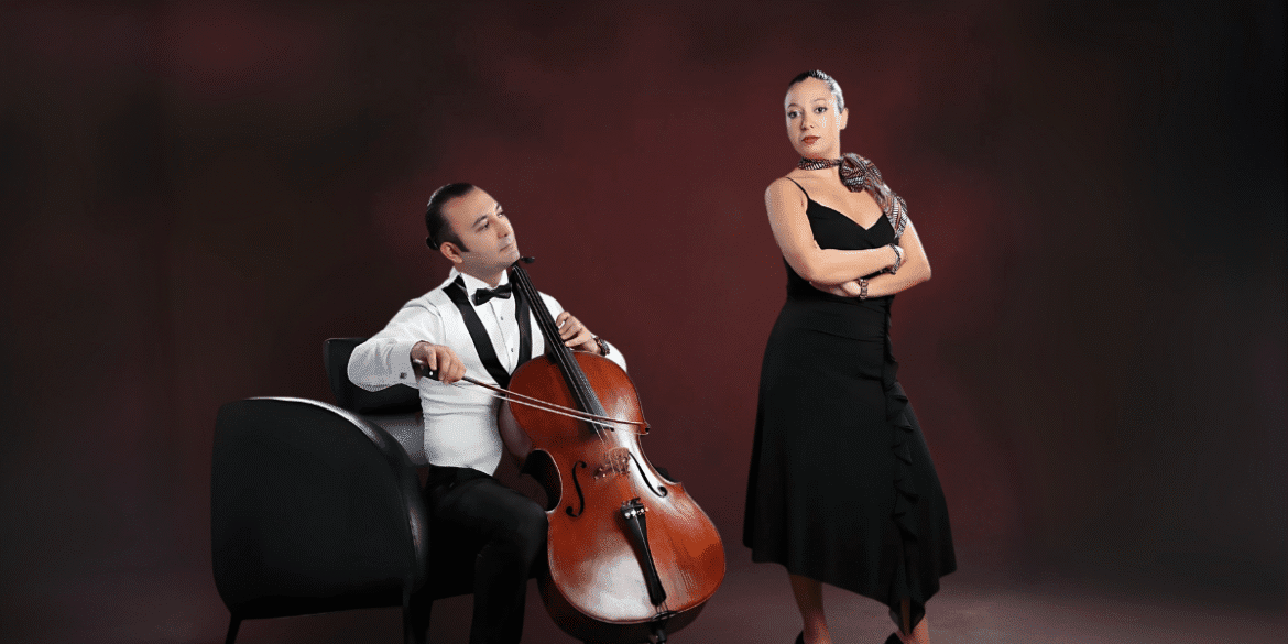 Man playing cello while woman in black dress stands beside him on stage.