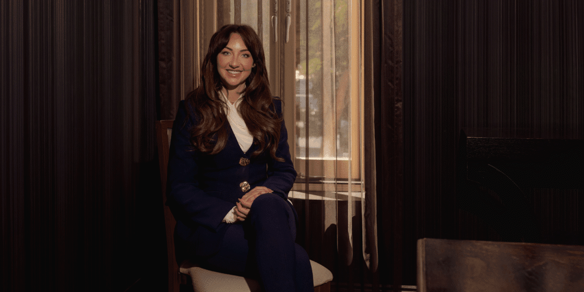 "Smiling woman in a blue suit sitting near a window, exuding confidence and warmth in a professional setting."