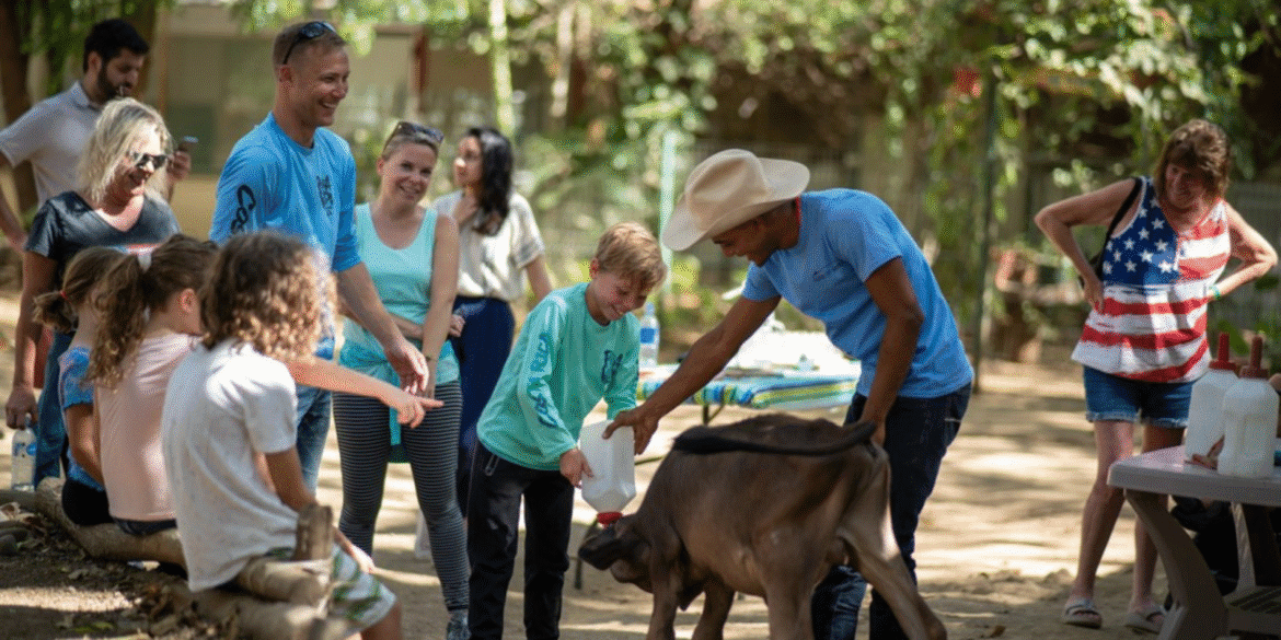 Children and adults smile while feeding a calf at The Monkey Farm’s community-focused recovery and healing center in Costa Rica.