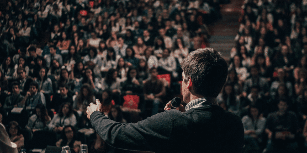 The image shows a speaker addressing a large audience at a conference or seminar, highlighting the importance of communication.