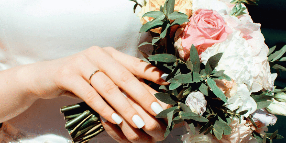 A close-up of a bride's hand holding a bouquet of pink and white flowers, showcasing her engagement ring and elegant manicure.