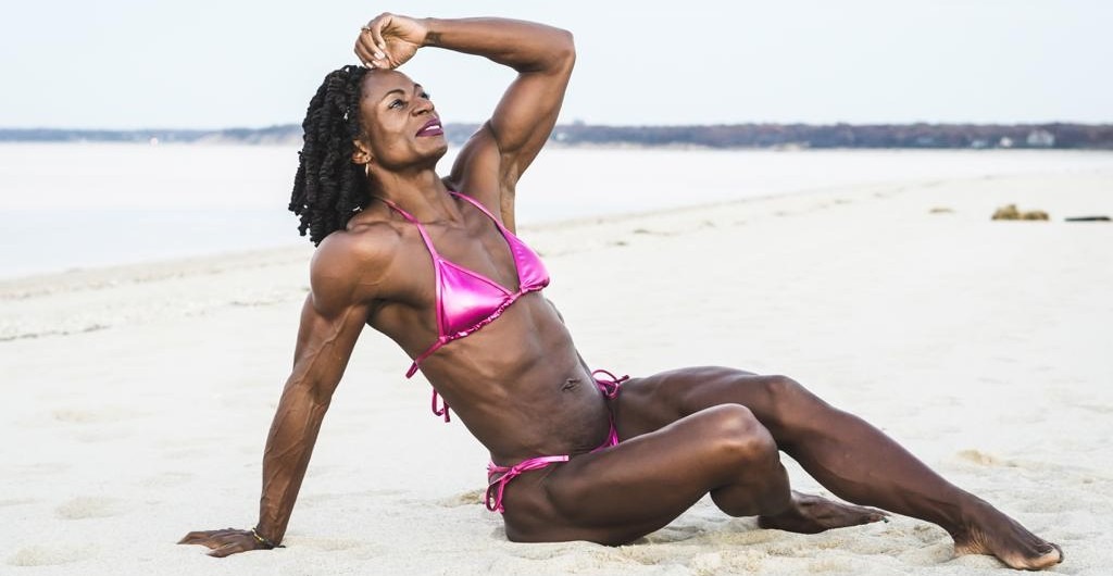 A strong, confident woman with muscular physique sits on a sandy beach, reflecting strength, discipline, and personal transformation.