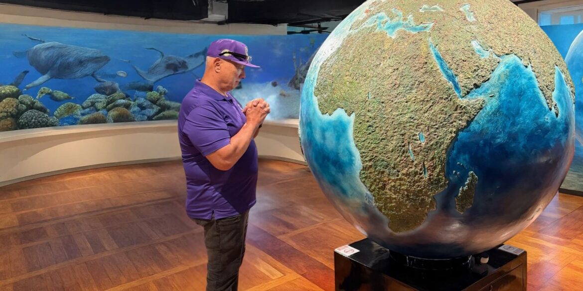 Apostle Constantine I. Nightingdale stands by a large globe, contemplating with hands clasped in a museum, surrounded by an underwater mural featuring humpback whales.