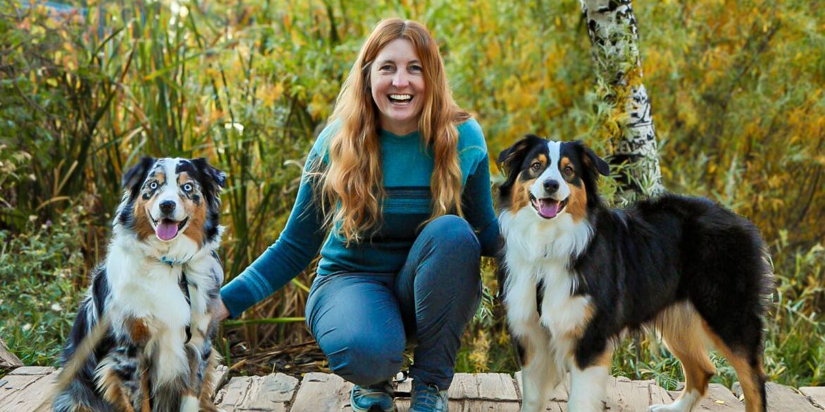 A woman smiles with two Australian Shepherd dogs in a natural setting, reflecting the bond built through expert training.