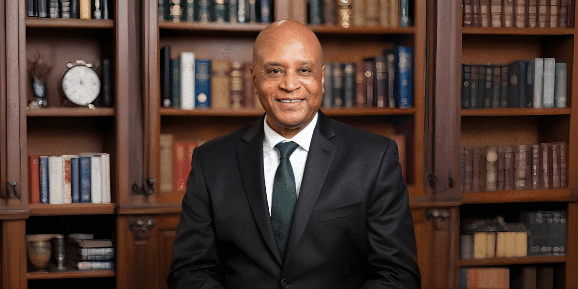 Smiling man in suit seated before wooden bookshelf filled with books in home office.
