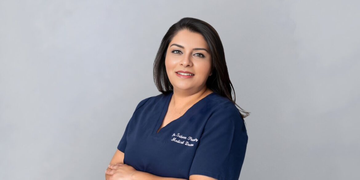 A woman in a blue medical uniform stands confidently against a gray background, smiling gently with arms crossed, conveying professionalism and warmth.