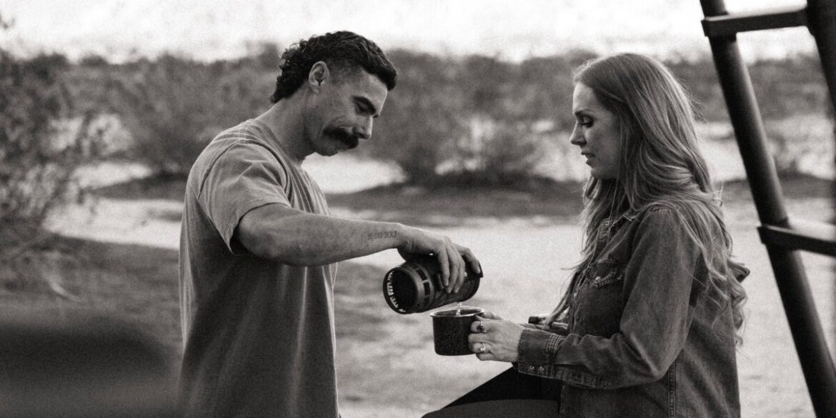 A man pours coffee into a mug held by a woman outdoors. The scene conveys a peaceful and warm moment, set against a backdrop of blurred natural scenery.