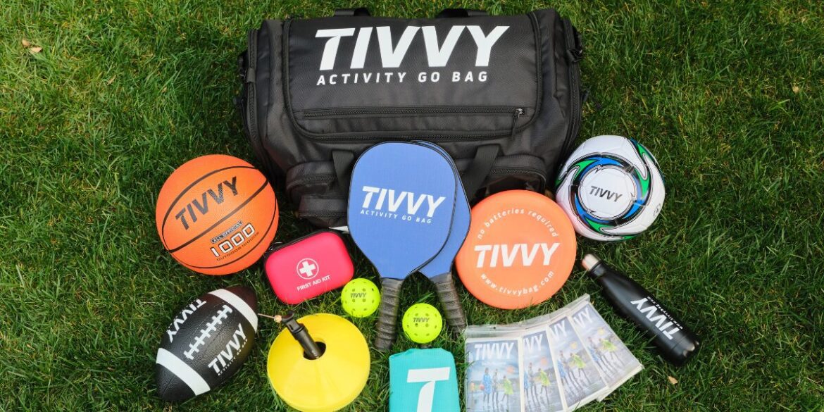 Black sports bag labeled "Tivvy" sits on grass surrounded by various activities gear: basketball, soccer ball, paddles, cones, water bottle, and discs.
