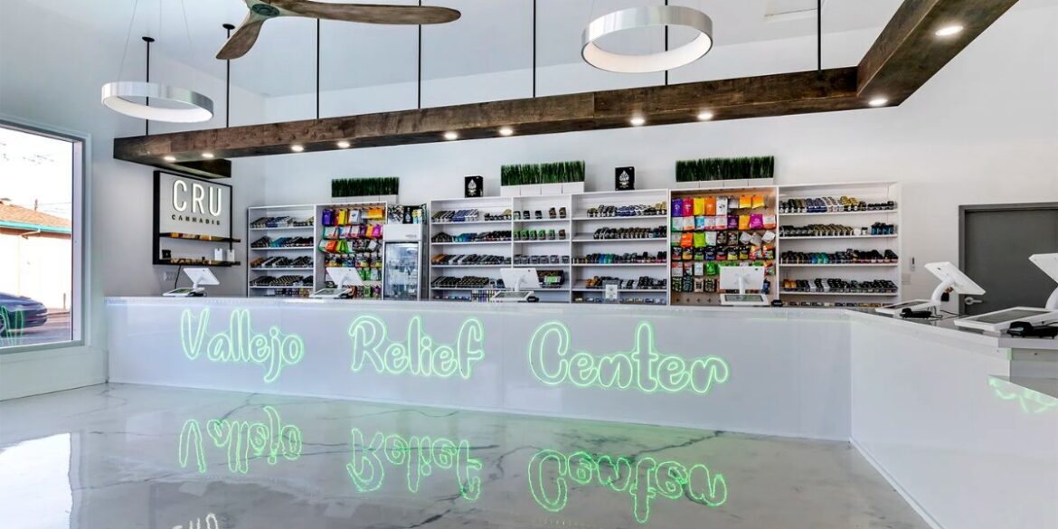 Spacious, modern dispensary interior with sleek white counters and shelves stocked with colorful products. Neon sign reads "Vallejo Relief Center."
