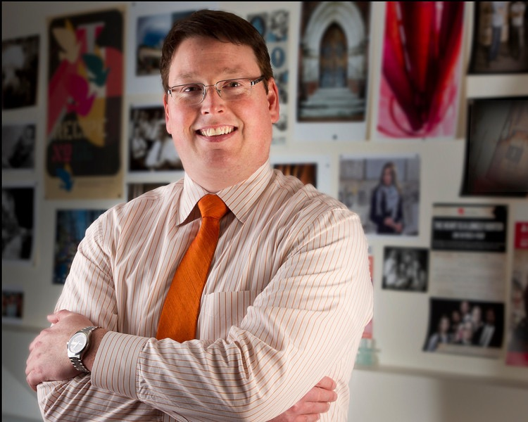 Nathan Hickman smiling with folded arms in front of a photo display.