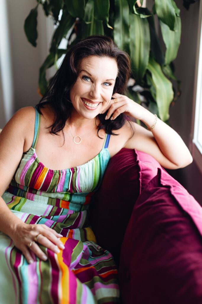 Katrina Foe smiling while seated on a sofa beside a large indoor plant.