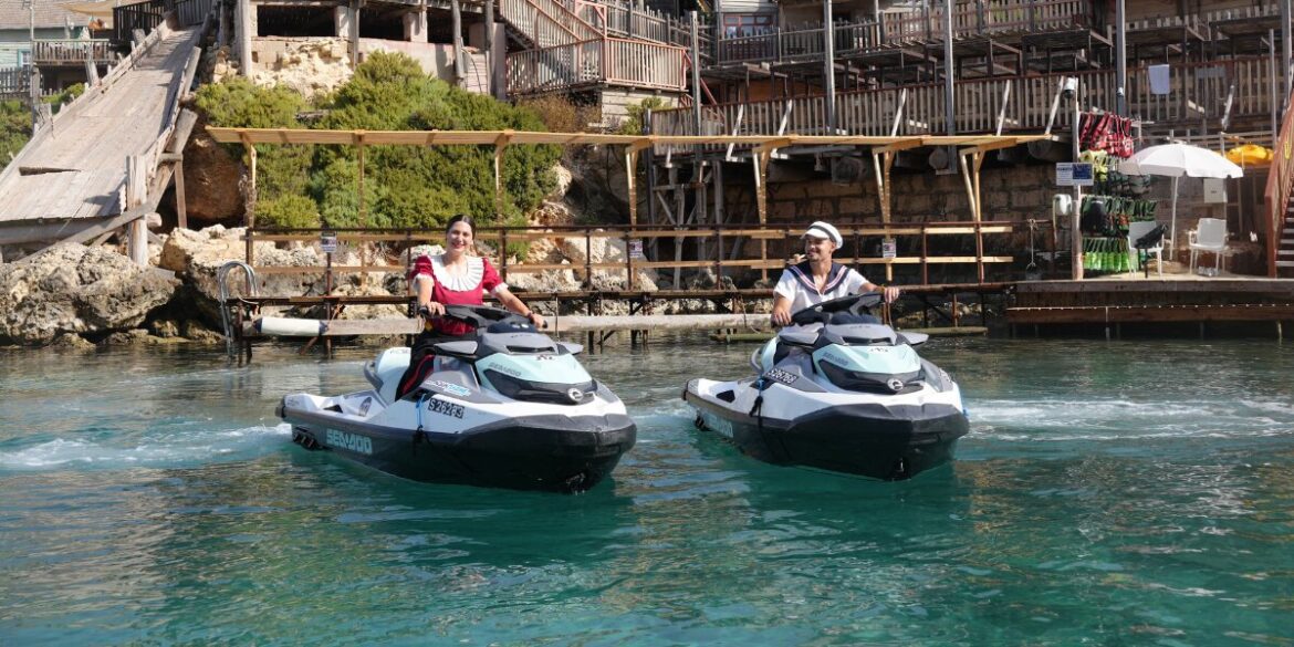 Two people ride Sea-Doo jet skis together on the water in front of Popeye Village.